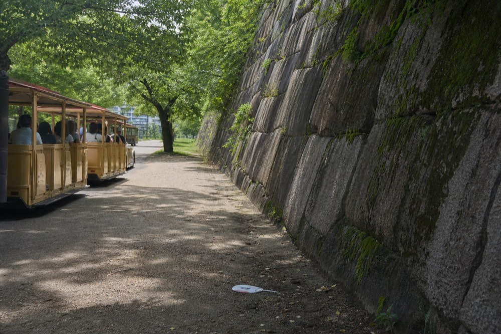 A green Road Train shuttle car passing in front of the stone walls of Osaka Castle
