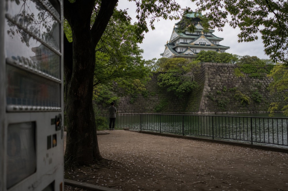 White-walled Osaka Castle tower with green roof tiles seen across the outer moat from the park path