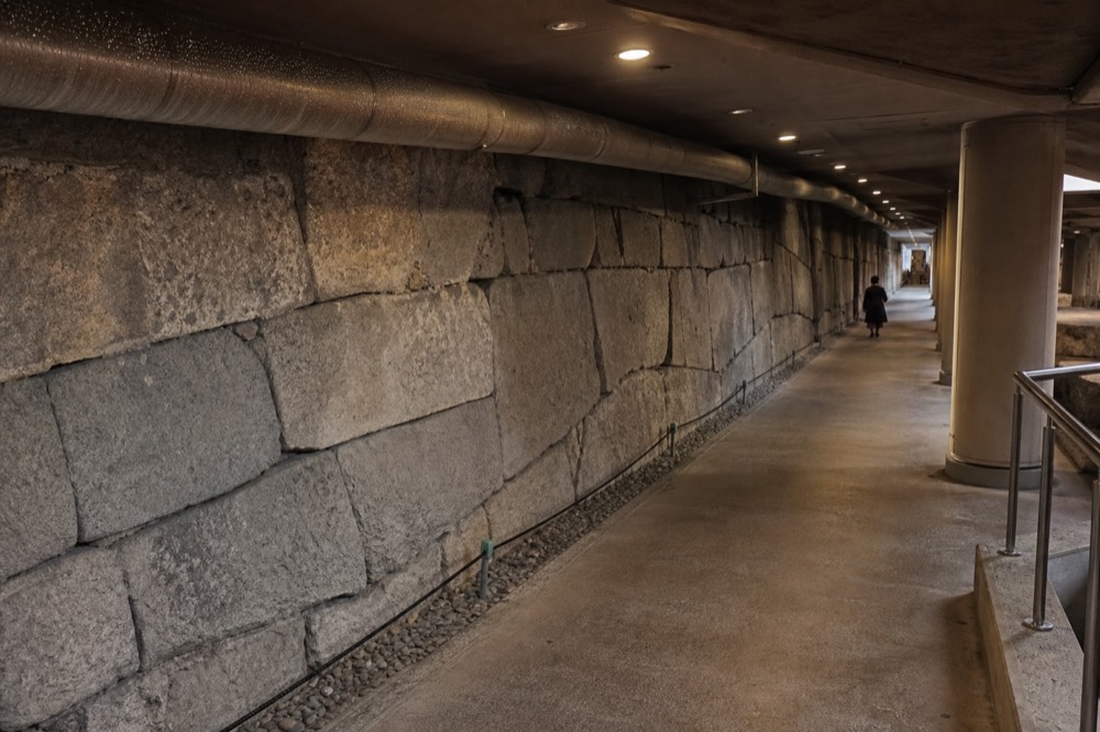 An underground exhibition hall showing an old stone wall lit behind a walkway