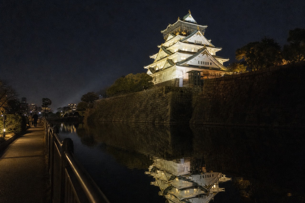 Osaka Castle Main Tower glowing gold against a dark sky, its reflection shimmering in the calm inner moat