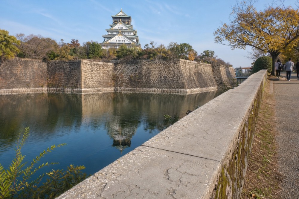 The gold-trimmed white tower of Osaka Castle seen across the inner moat from the stone wall
