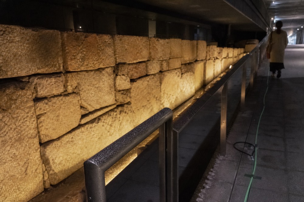 An underground exhibition hall with an ancient stone wall lit along its length beside a walkway